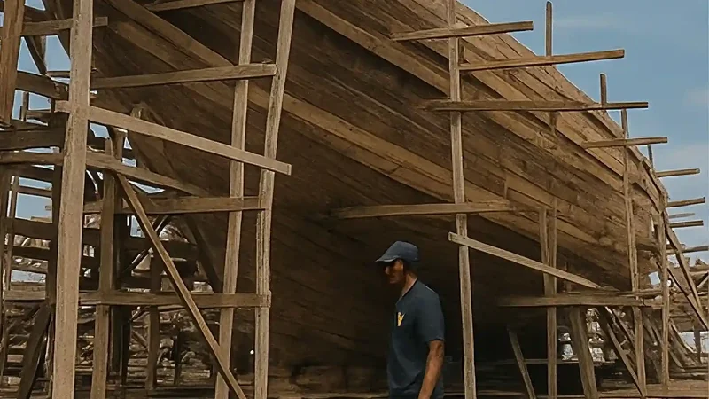 Large wooden hull enclosed in bamboo scaffolding, a single shipwright standing beside it for scale