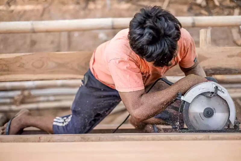 Shipwright running a circular saw along a long timber plank on the yard floor