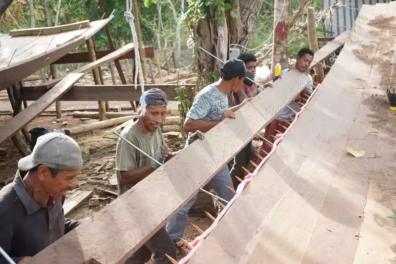 Several shipwrights working together along a single long plank at the yard