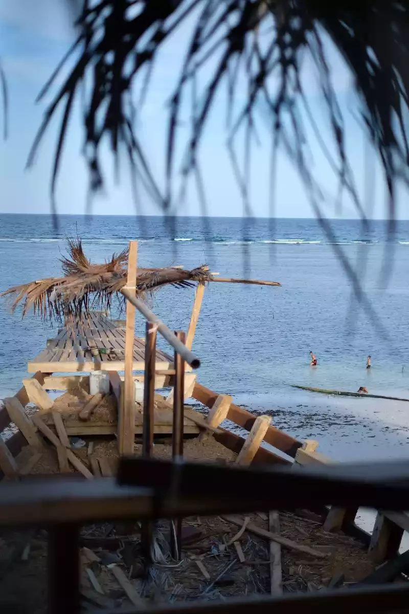 Wooden hull frames standing under a palm-thatch shelter at the edge of the beach