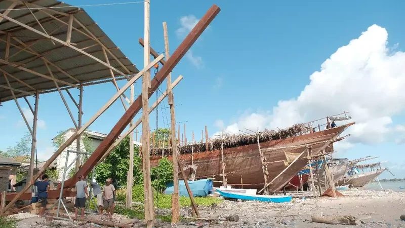 Wooden phinisi hull on timber props under a palm-thatch shelter at the water’s edge