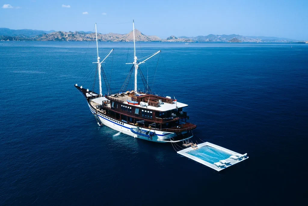 Wooden phinisi lying at anchor in calm water off a green headland