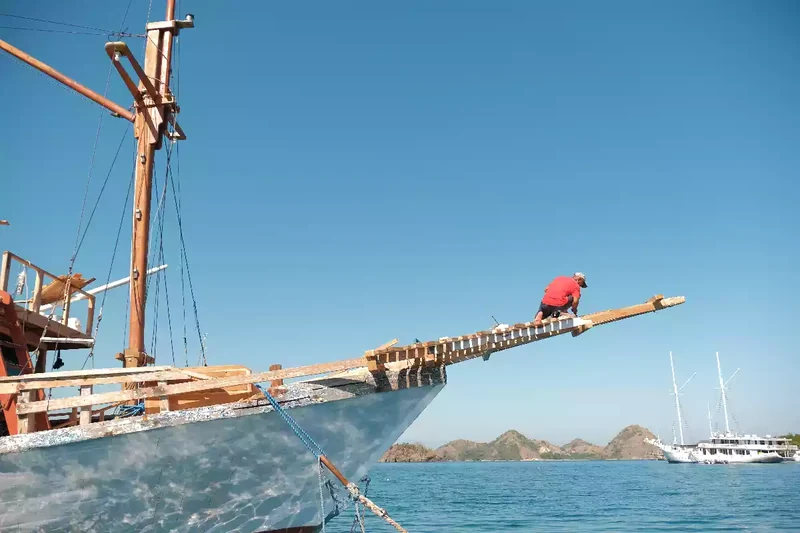 Crew member working out along the bowsprit of a wooden phinisi, masts and standing rigging behind