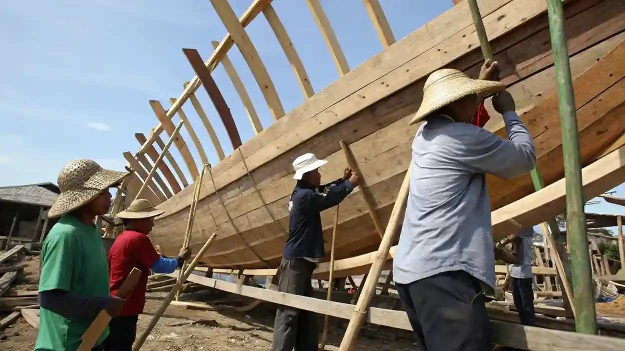 Shipwrights in sun hats working along the frames of a wooden hull propped on the sand