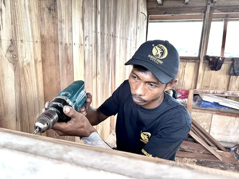 Tukang kapal sedang mengebor dan mengerjakan papan kayu pada lambung kapal yang dibangun