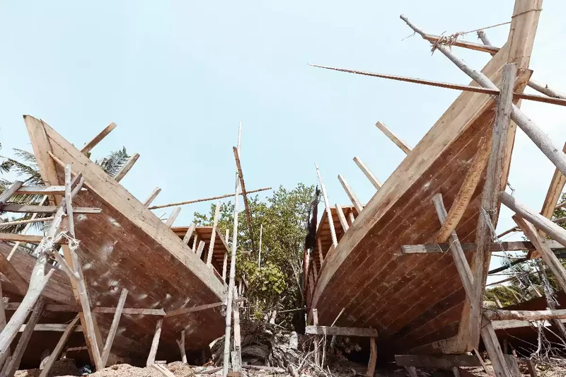 Two wooden phinisi hulls standing on timber props at a beach boatyard, planking part-finished
